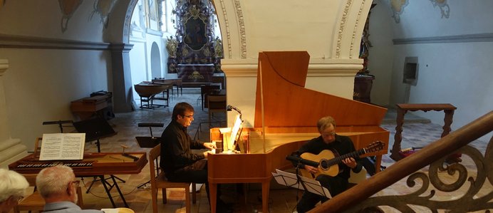 Ein Kirchenraum in welchem eine Person an einem Flügel spielt und eine weitere Person mit eine akustischen Gitarre. 