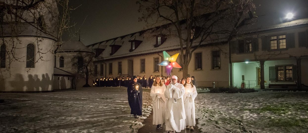 Eine Gruppe Sternsingerinnen und Sternsinger vor der Klosterkirche Wettingen. Die vordersten Sänger (in weiss) halten einen leuchtenden Stern in die Höhe. Am Boden liegt Schnee.