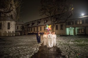 Eine Gruppe Sternsingerinnen und Sternsinger vor der Klosterkirche Wettingen. Die vordersten Sänger (in weiss) halten einen leuchtenden Stern in die Höhe. Am Boden liegt Schnee.