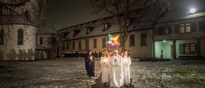 Eine Gruppe Sternsingerinnen und Sternsinger vor der Klosterkirche Wettingen. Die vordersten Sänger (in weiss) halten einen leuchtenden Stern in die Höhe. Am Boden liegt Schnee.